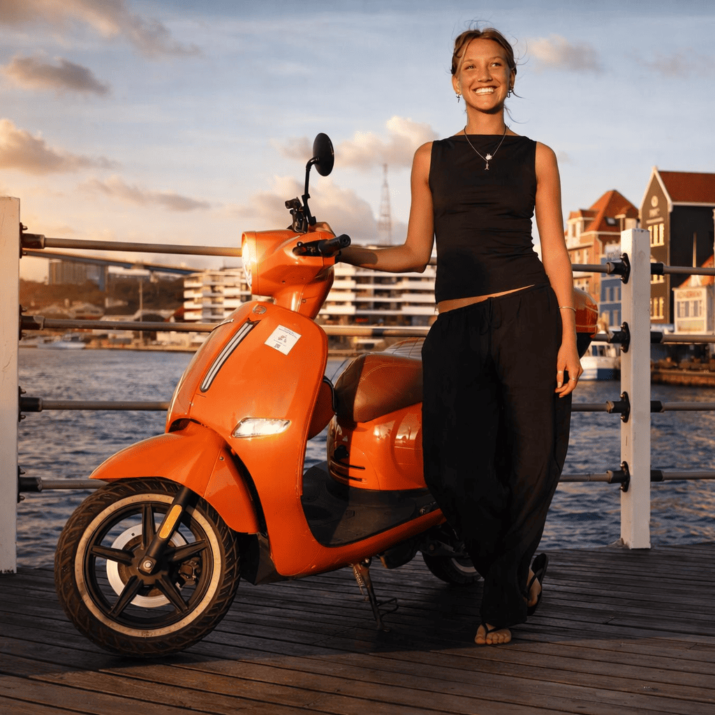 Female student standing next to orange Flamingo Go scooter near waterfront in Curaçao